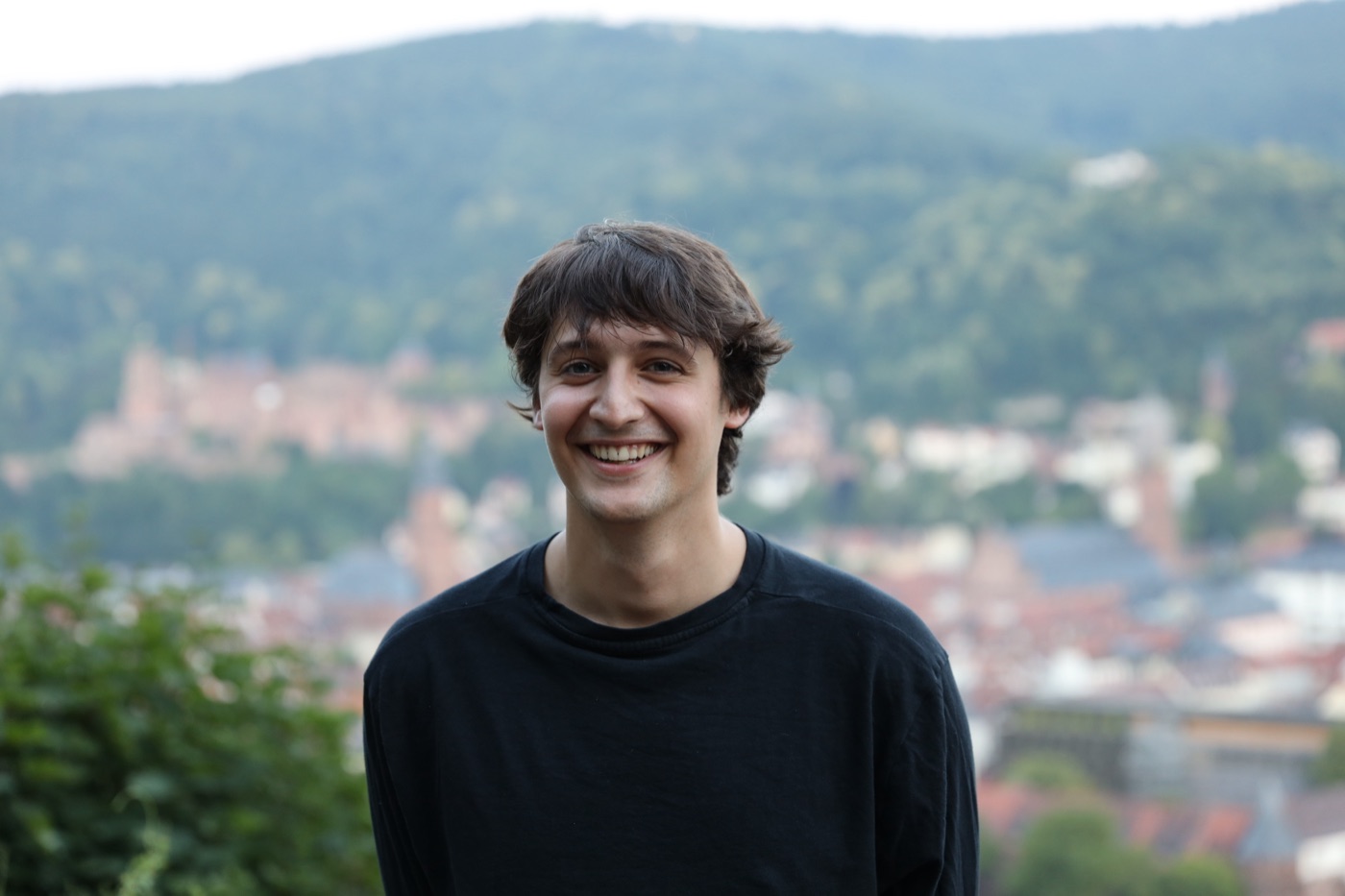 Tiago, smiling, in a black jumper, with a town and wooded hills out of focus behind him.